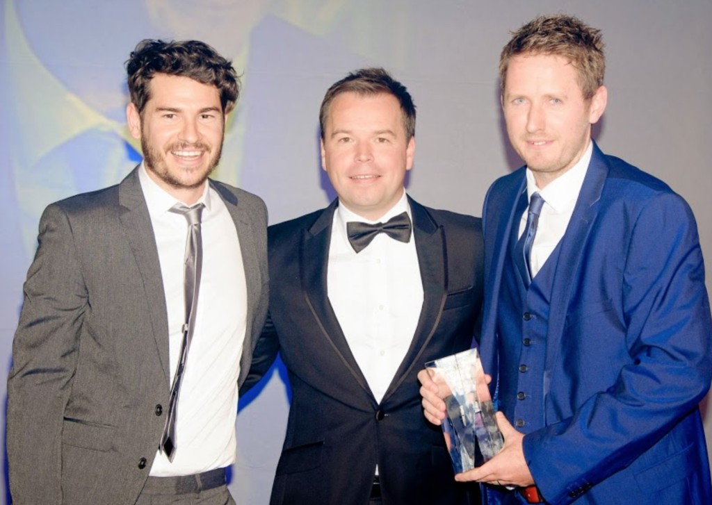 Three men standing looking at camera, one in a grey suit, one in black and one in a blue suit, holding an award